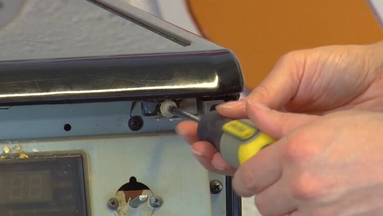 Use a Phillips screwdriver to remove the two screws that hold the top panel in place at the front of the oven