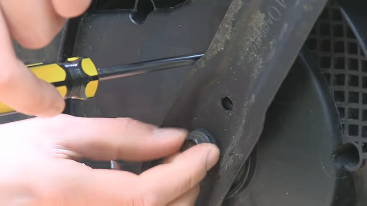 Placing A Screwdriver In The Hole Above Lawnmower Blade While Unscrewing The Central Locking Nut By Hand