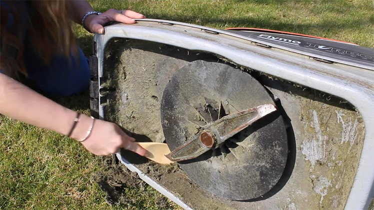 Scraping Away Any Caked On Grass From The Lawnmower Surface And The Blades With A Wooden Spoon